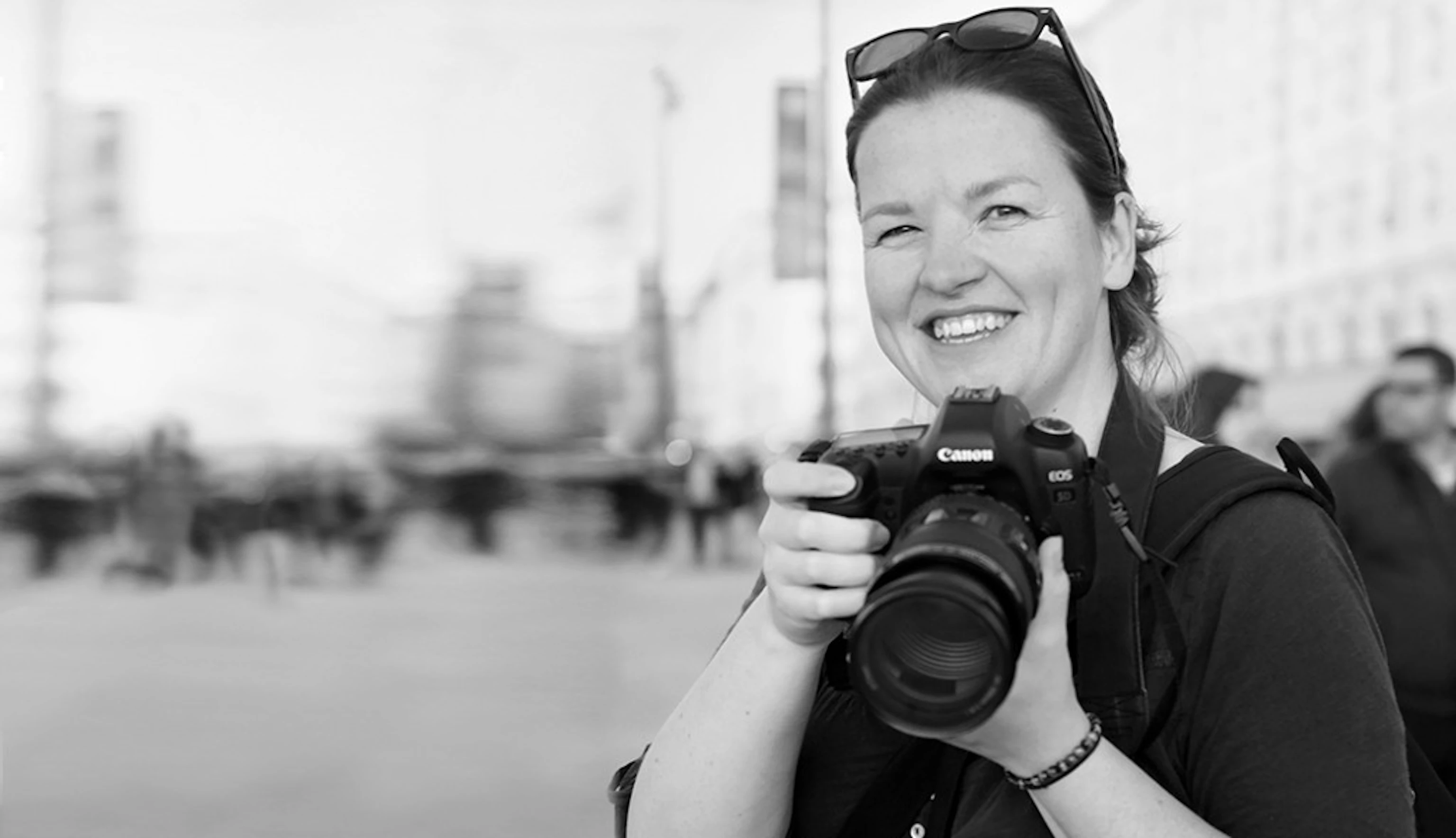 Rachel Bywater stands to the right smiling into the camera. She is holding a camera with both hands just below her face. A background of passersby is out of focus.