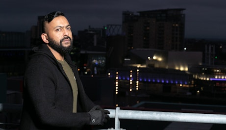 Sam Malik standing side on leant against a metal bar looking into the camera. Behind him is a nighttime Manchester city skyline landscape.
