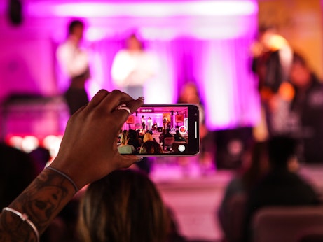 A hand holding a phone up and recording women on a pink stage. The stage itself in front is blurred, with focus on the hand and the phone. The scene in front can be seen clearly through the phone camera.