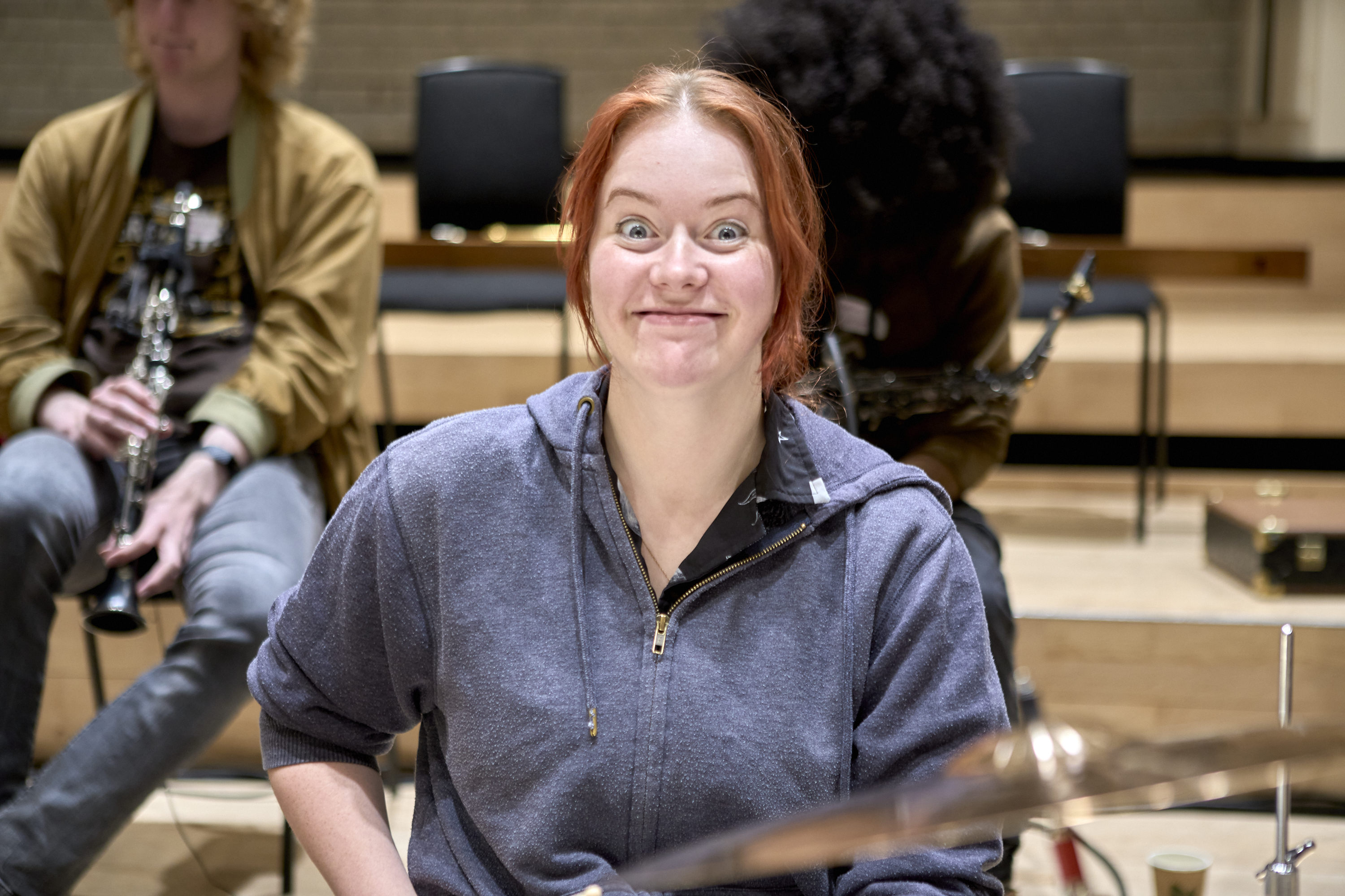 A young musician sat at a drum kit smiling widely into the camera.