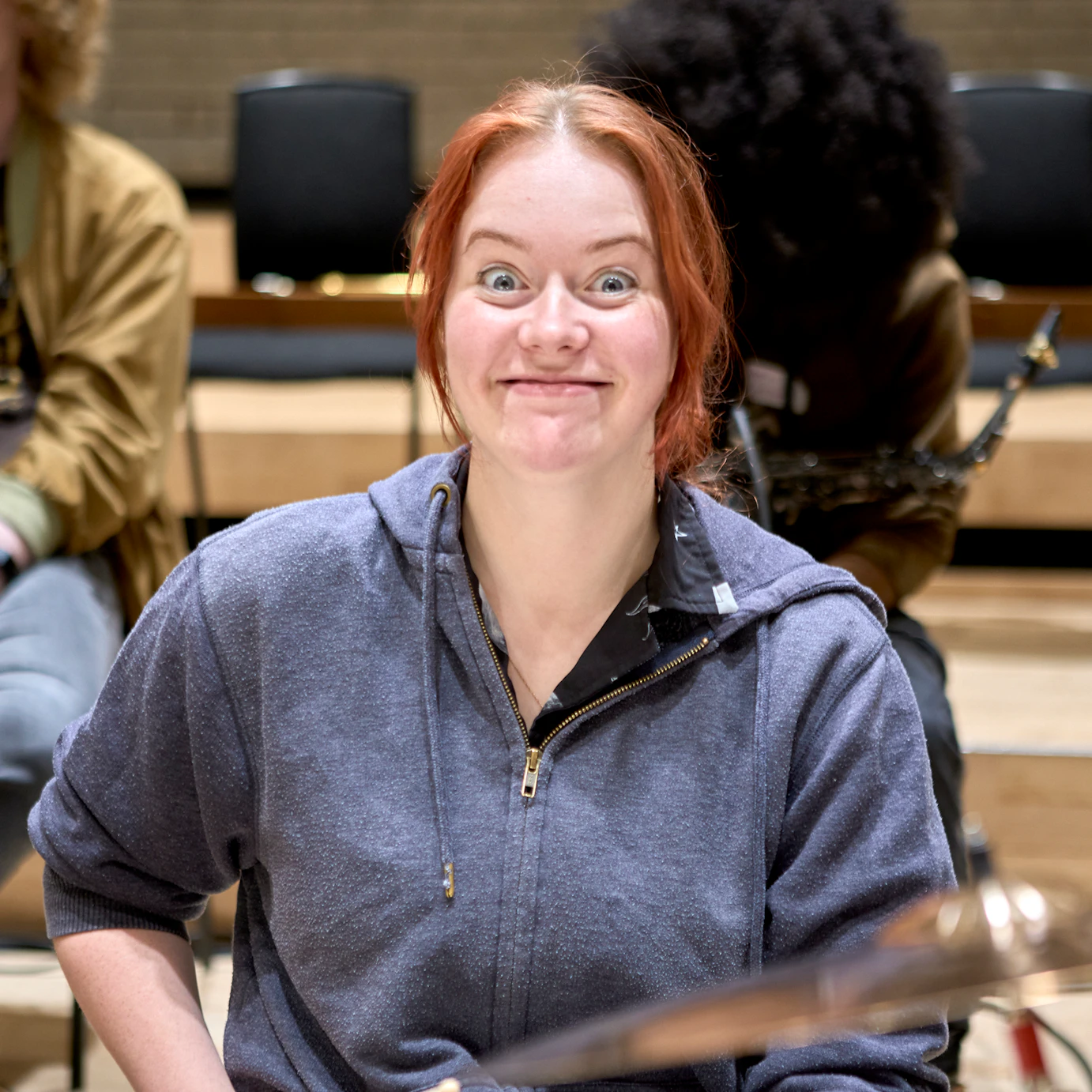 A young musician sat at a drum kit smiling widely into the camera.