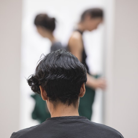 Fatima Al Qadiri from behind looking on at two blurred residency artists in front stood back to back between two white monoliths.