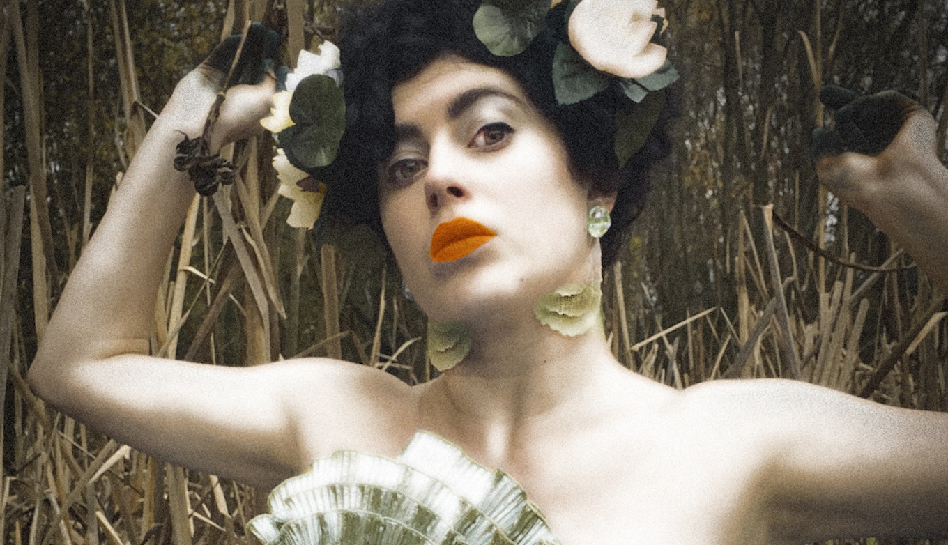 Ruby Tingle wears a light green dress with flowers in her hair stood amongst tall grass and trees. Her hands are black, possibly caked in mud.