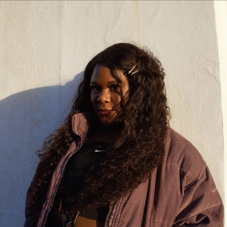 Artist Ifeoluwa standing against a white wall looking into the camera.