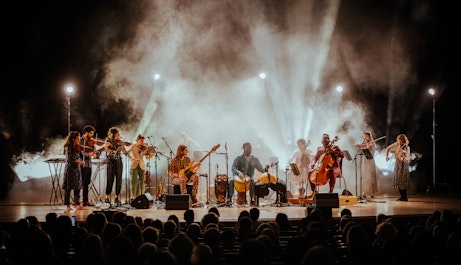A group of ten musicians playing different instruments play on a stage to a large audience.