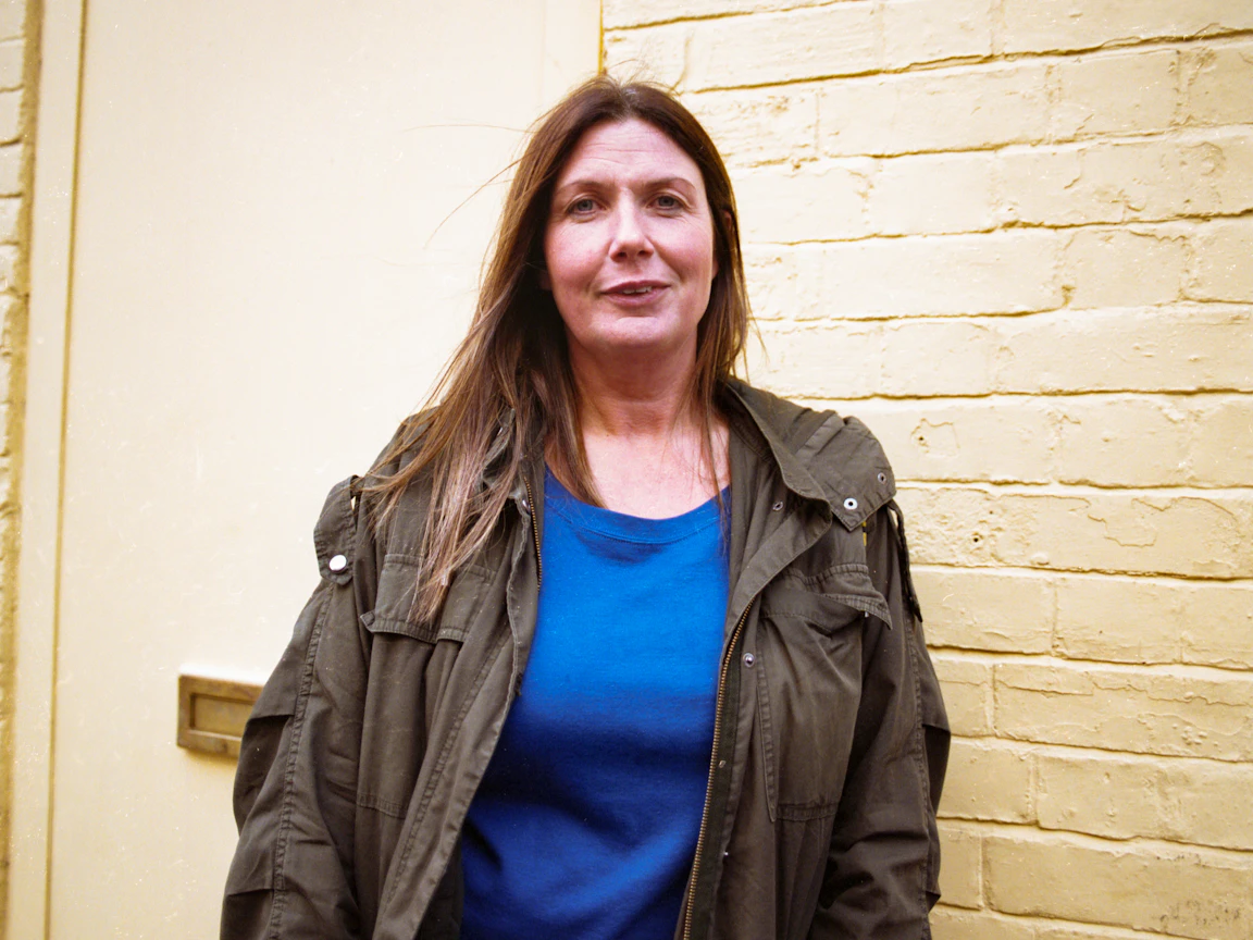 Kate smiles at the camera. Kate is wearing a blue t-shirt and khaki coat whilst standing in front of a yellow brick wall.