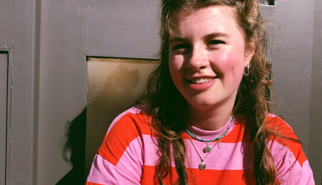 Claire Hamilton smiling off camera sat in front of a door and wearing a stripy red and pink shirt.