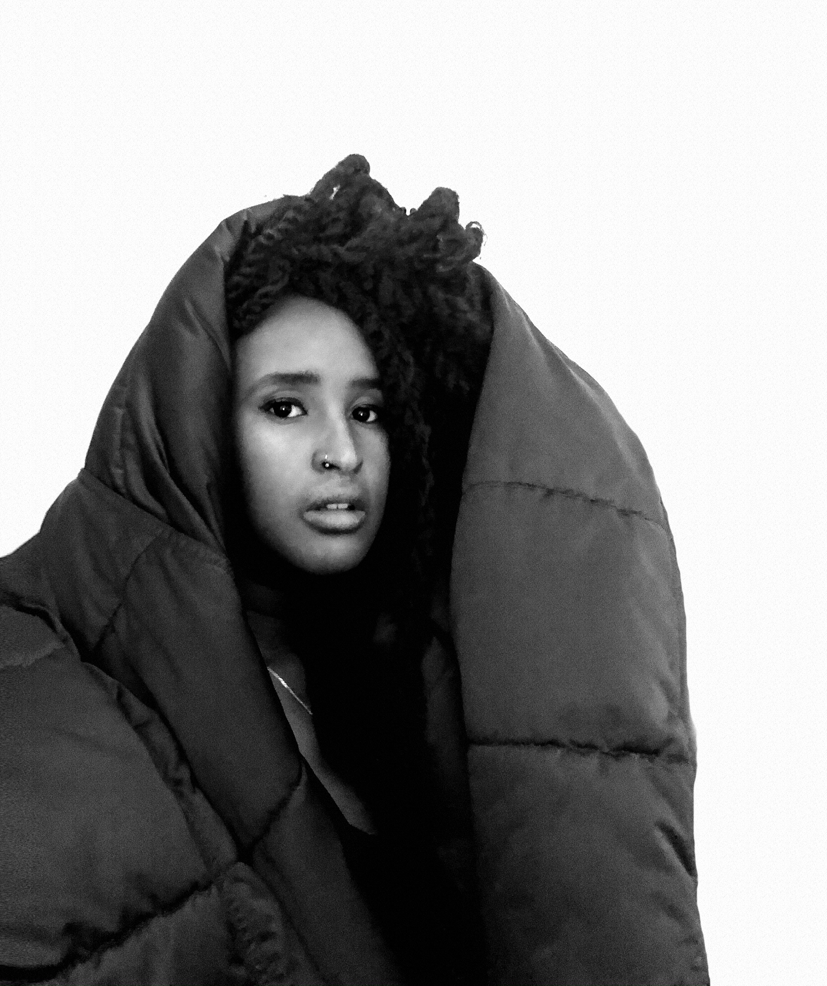 Hur̃guf on a white background looling into the camera. Over her head and shoulders is what looks like an oversized puffer coat or sleeping bag.