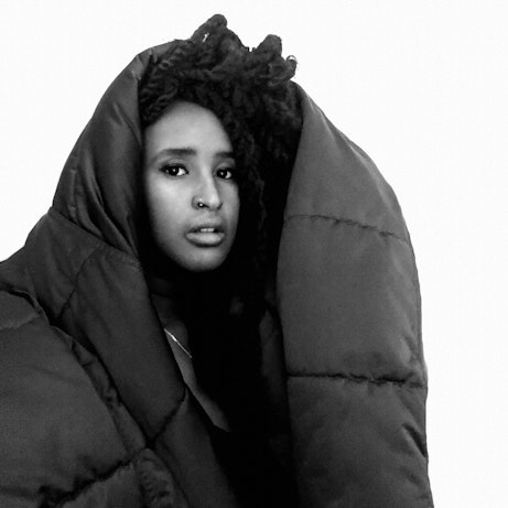 Hur̃guf on a white background looling into the camera. Over her head and shoulders is what looks like an oversized puffer coat or sleeping bag.
