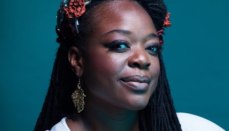 Headshot of ESKA smiling at the camera with flowers in her hair against a teal background.