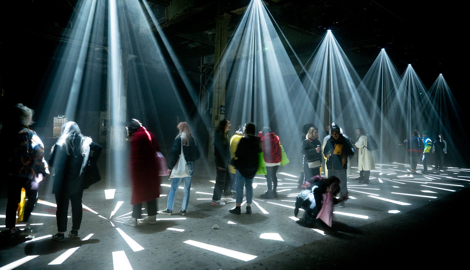 A group of fifteen people are stood under projections of light in a dark space.