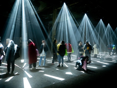 A group of people stand under strobe projections from the ceiling. The projections are making different shapes on the floor. Some people are looking up at the ceiling.