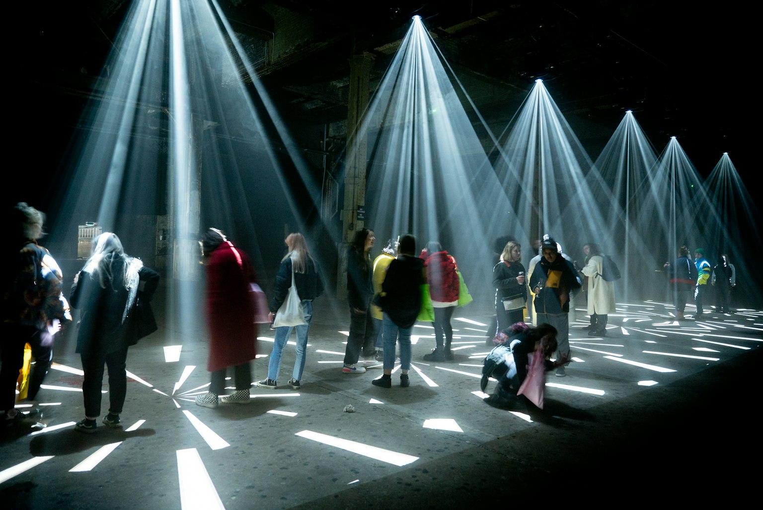 A group of people stand under strobe projections from the ceiling. The projections are making different shapes on the floor. Some people are looking up at the ceiling.