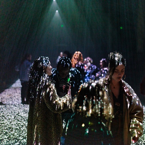 In a dark space, various people are illuminated in dots of colour from projections shining down from above. The dots of light cover the people and the floor around them.