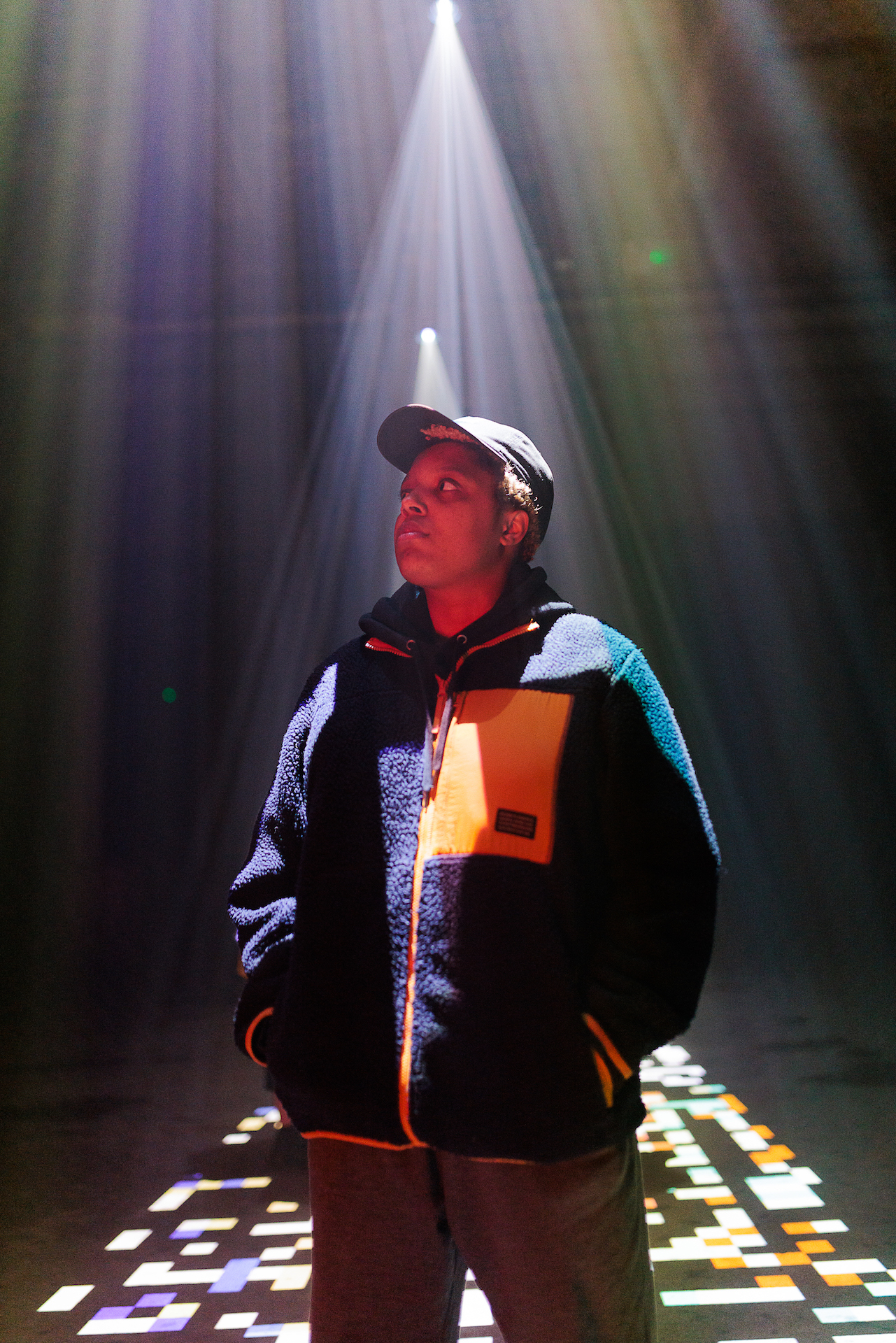 Loraine James stood underneath projections shining down from the ceiling. She has her hands in her coat pockets and is looking off to her right. On the floor around her are projections of disjointed pixels in various colours.