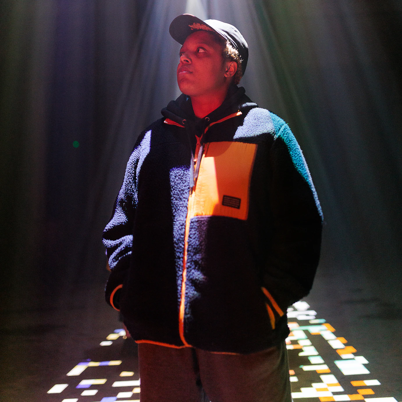 Loraine James stood underneath projections shining down from the ceiling. She has her hands in her coat pockets and is looking off to her right. On the floor around her are projections of disjointed pixels in various colours.