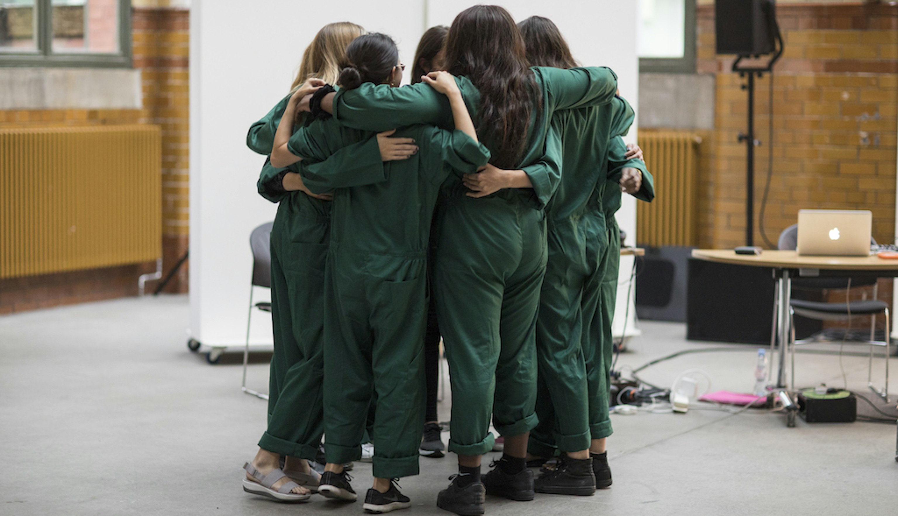 A group hug. Everyone is wearing dark green boiler suits with arms around one another's shoulders.