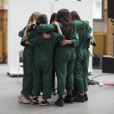 A group hug. Everyone is wearing dark green boiler suits with arms around one another's shoulders.