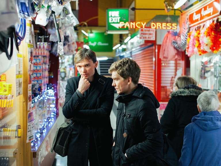 Field Music in black coats looking pensive. They are in an indoor market surrounded by products, bright neon signs and shutters.