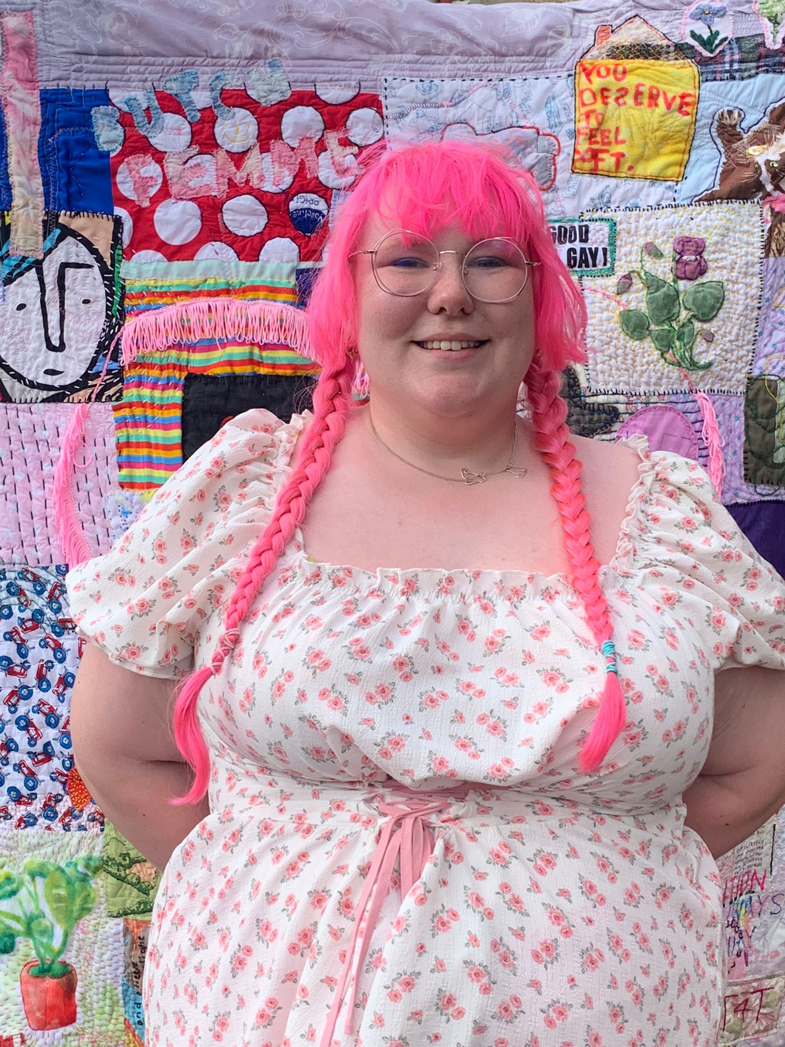 Eleanor Louise West stood smiling into the camera with their arms behind their back. They have pink pigtails in and are wearing a white dress with pinks flowers on. Behind is a knitted blanket with multiple patches stitched together, all with different images and colours.