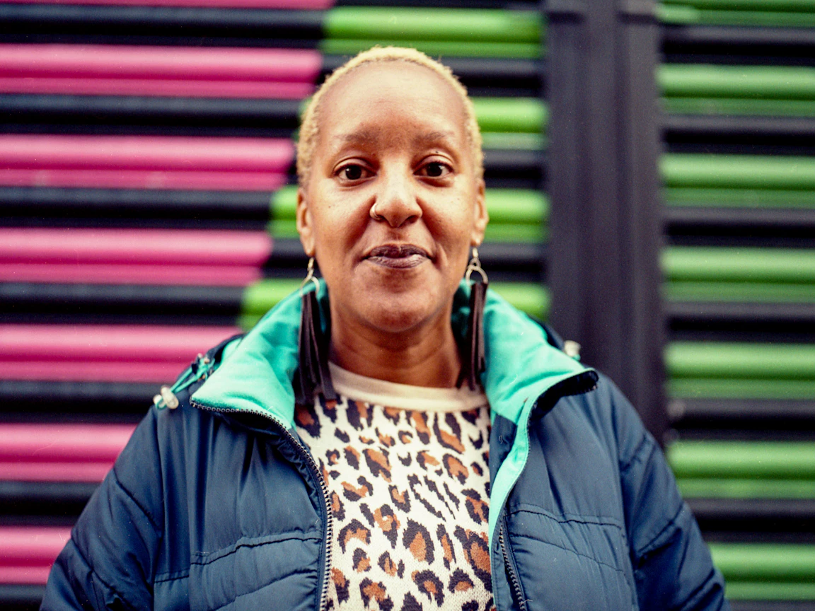Andrea is wearing a blue coat and is smiling at the camera. Andrea stands in front of green and pink shutters.
