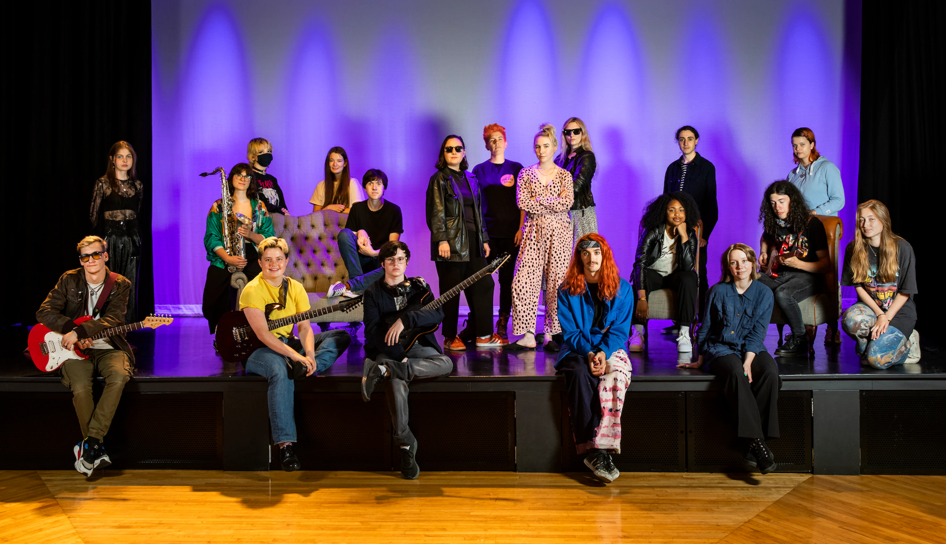 The band PINS stand on stage. They are surrounded by the participants from the project. Everyone is looking at the camera, some are smiling and some are holding instruments.