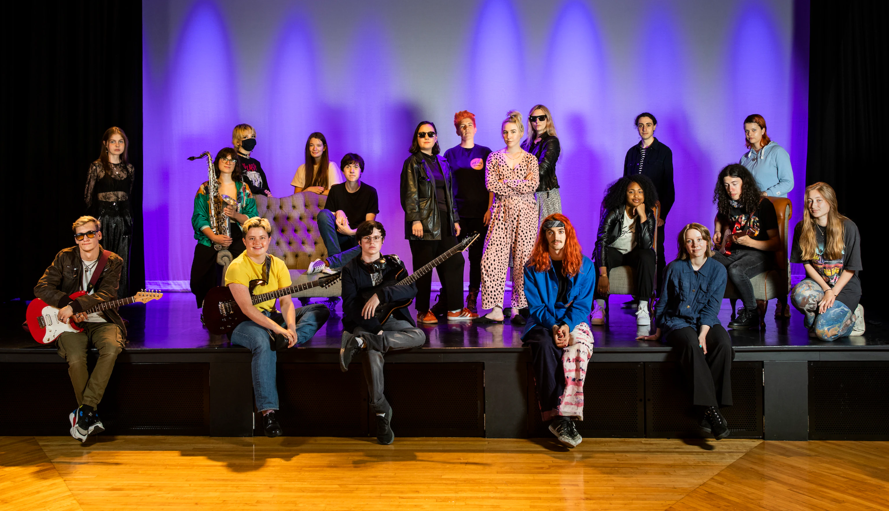 The band PINS stand on stage. They are surrounded by the participants from the project. Everyone is looking at the camera, some are smiling and some are holding instruments.