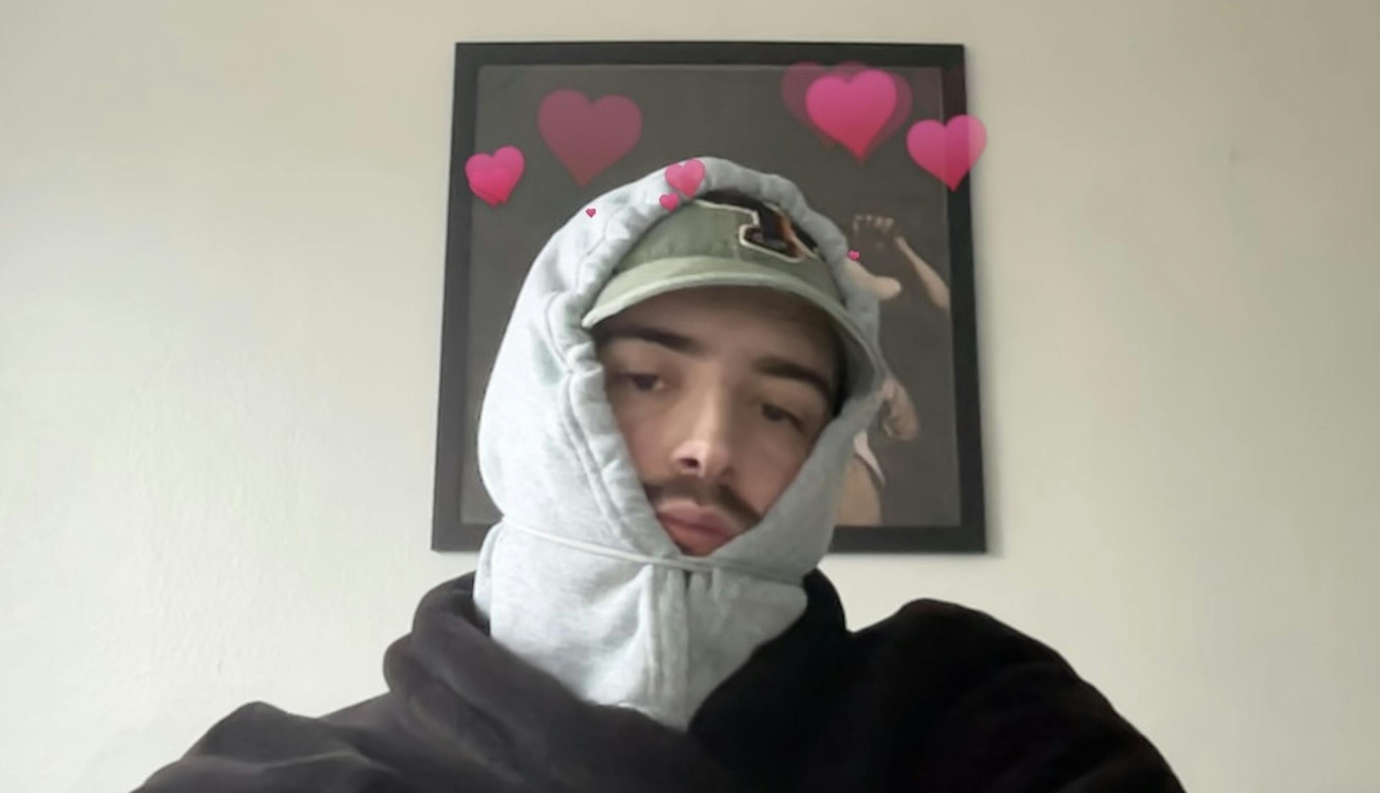 Louis Corts wearing a cap with a hoodie over it. Around his head are varying sizes of pink heart emojis.