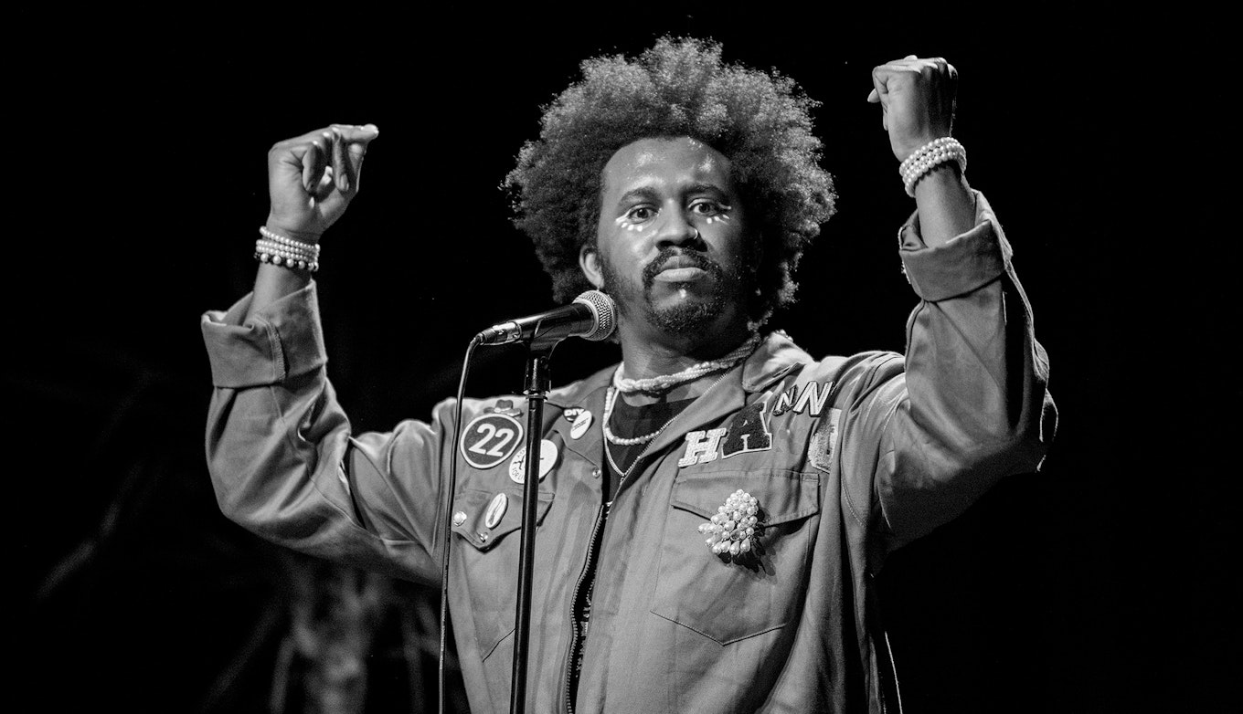 A black and grey image of Hang Linton performing. He's wearing a boiler suit with various badges and his arms are both raised halfway.