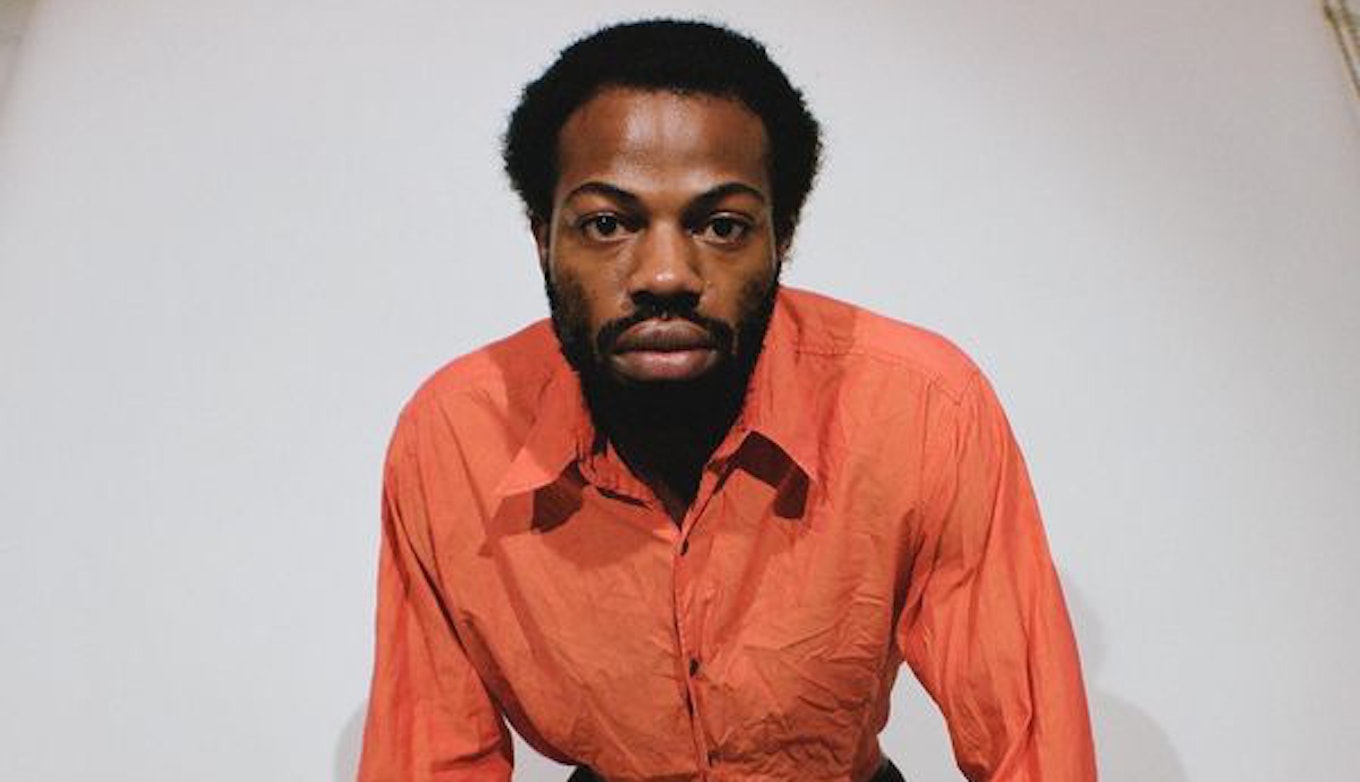 Ku-Ro bent with his hands on his knees looking into the camera. He's wearing a long-sleeve orange shirt and black trousers on a white wall background.