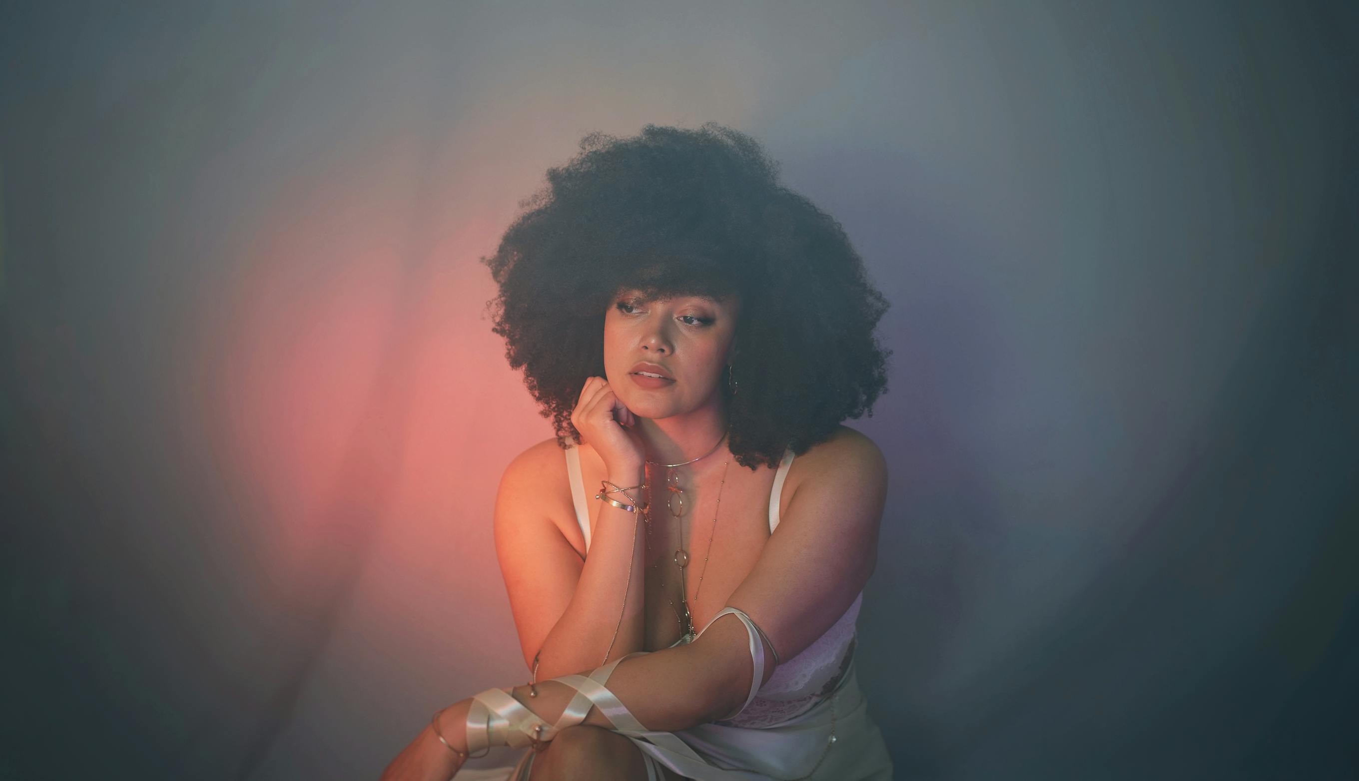 Elle Chante is crouched in front of a dark grey background, lit slightly by a warm red/peach light.