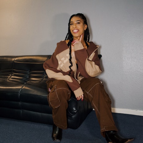 Victoria Jane dressed in light and dark brown baggy clothes with black heels. She is perched on the edge of a black sofa and smiling.