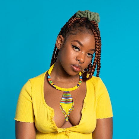 Meduulla is looking into the camera, wearing a yellow top and standing in front of a light blue background. She is wearing a beaded necklace that is primarily yellow with various other colours dotted throughout.