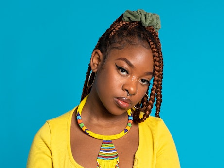 Meduulla is looking into the camera, wearing a yellow top and standing in front of a light blue background. She is wearing a beaded necklace that is primarily yellow with various other colours dotted throughout.