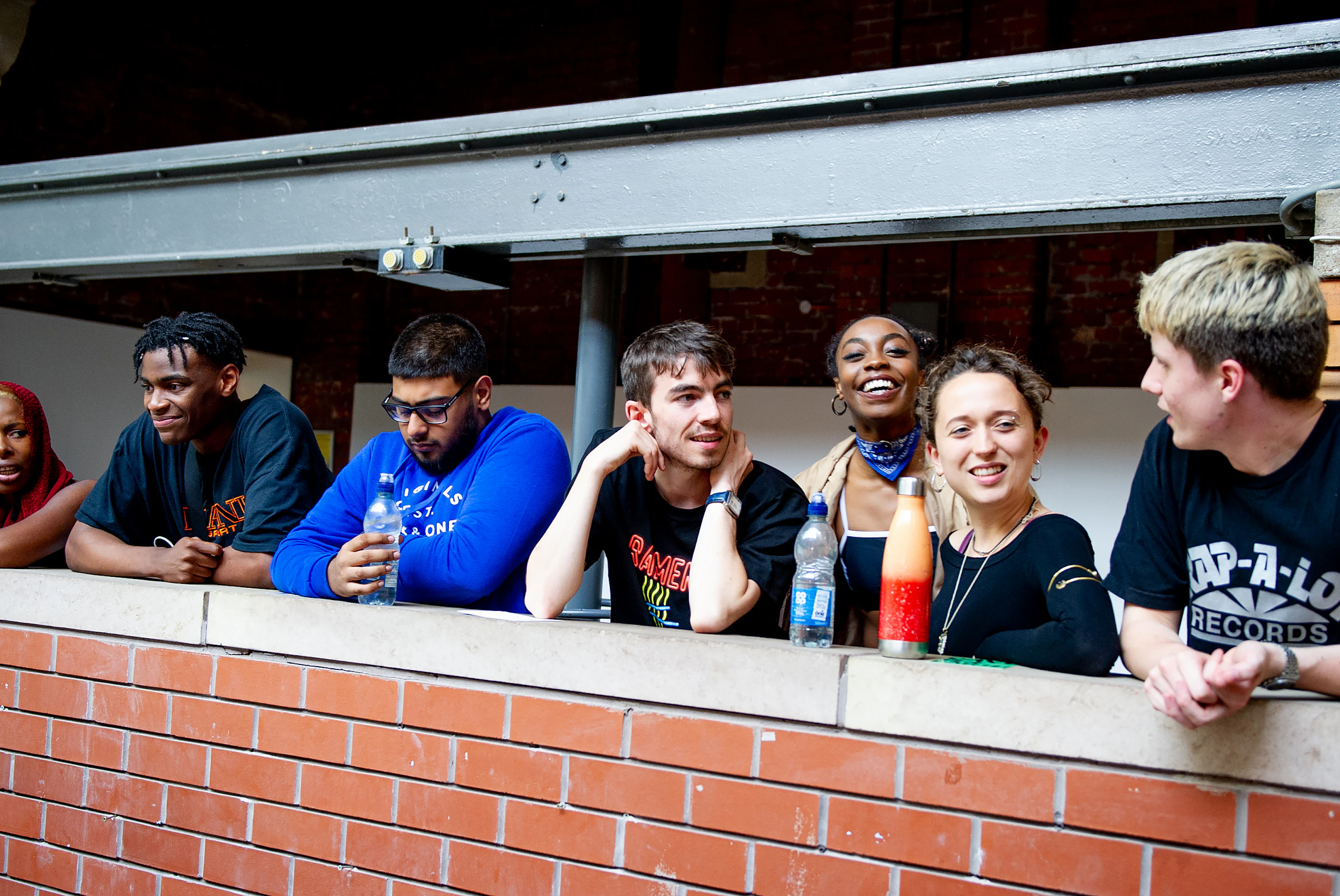 Young people stood next to one another leaning on a brick wall. Some are smiling at one another, others straight to camera.