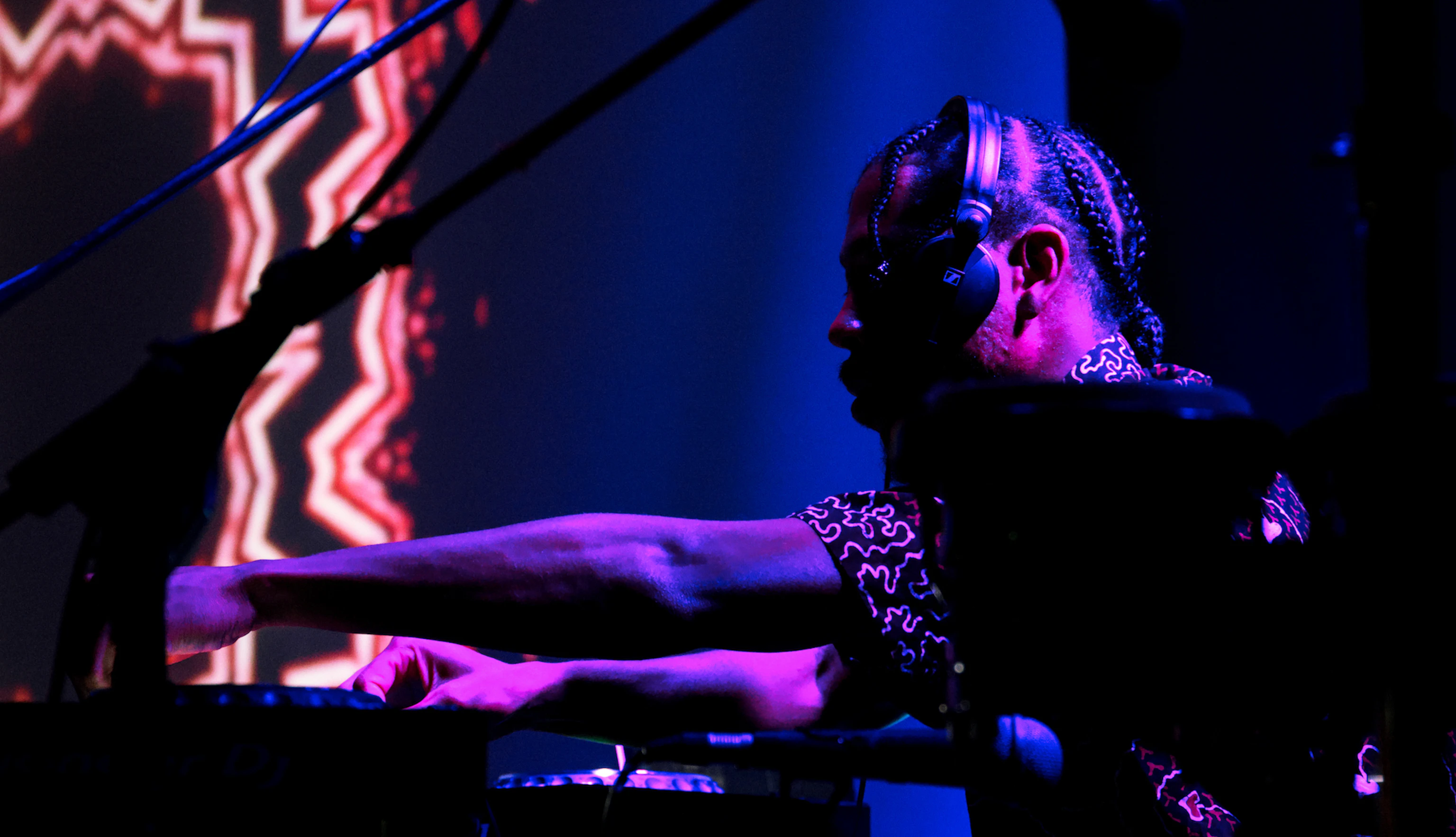 A musician with headphones on lit by purple and pink lights. In the background are projections of visual art.