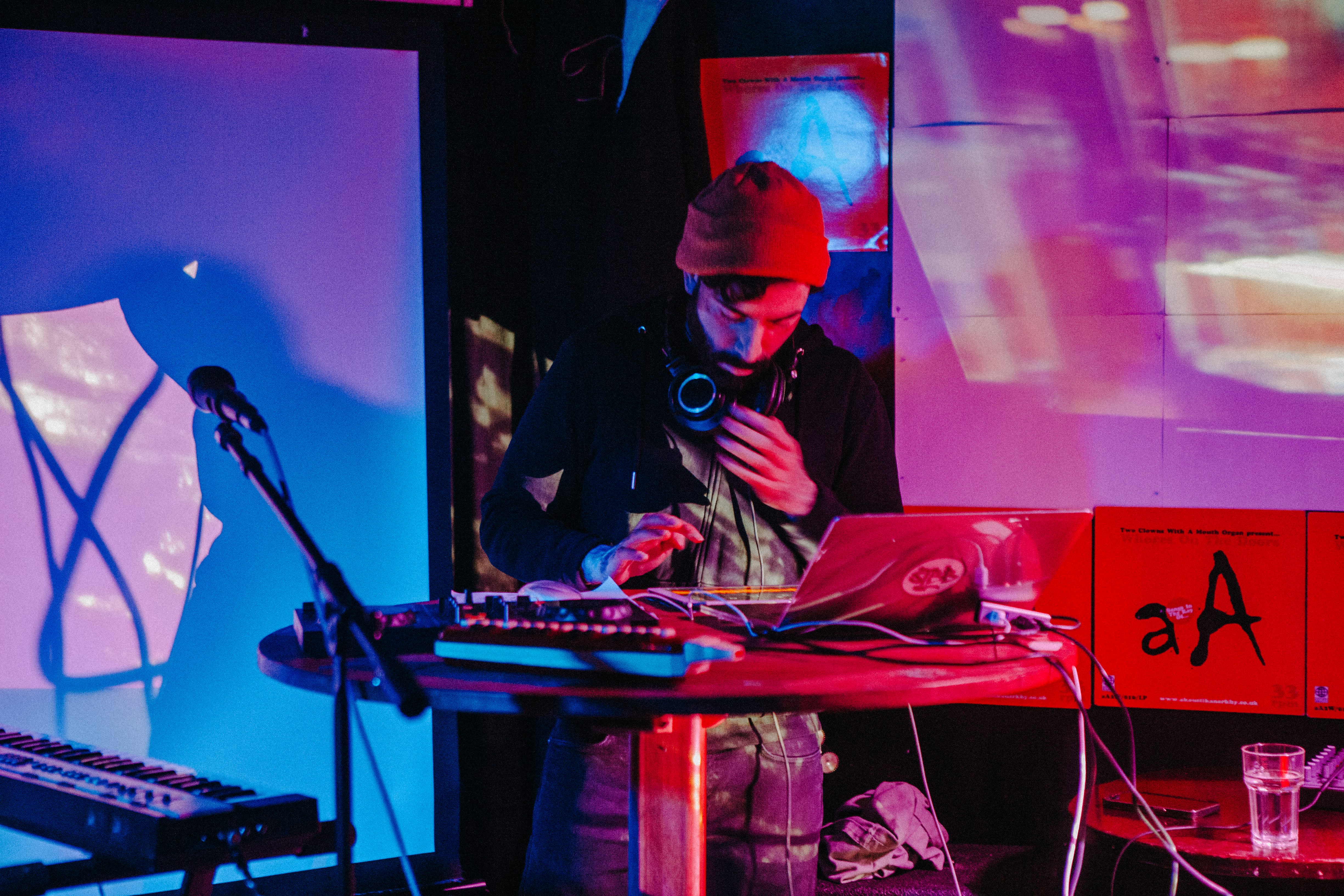 Astrolayb on stage behind a table with a laptop and various electronic musical equipment around him. Behind him are various projections lit by red and blue lighting.