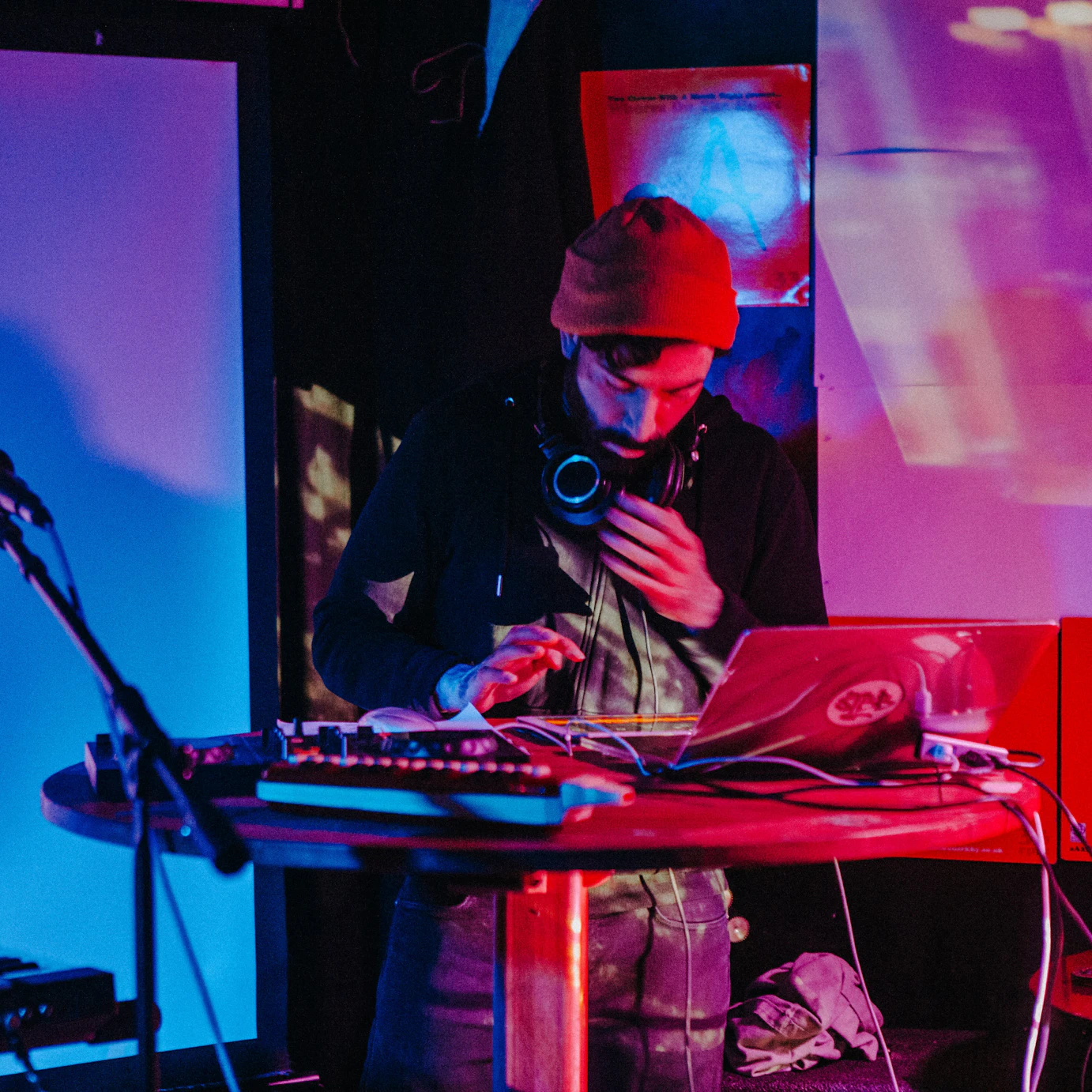 Astrolayb on stage behind a table with a laptop and various electronic musical equipment around him. Behind him are various projections lit by red and blue lighting.