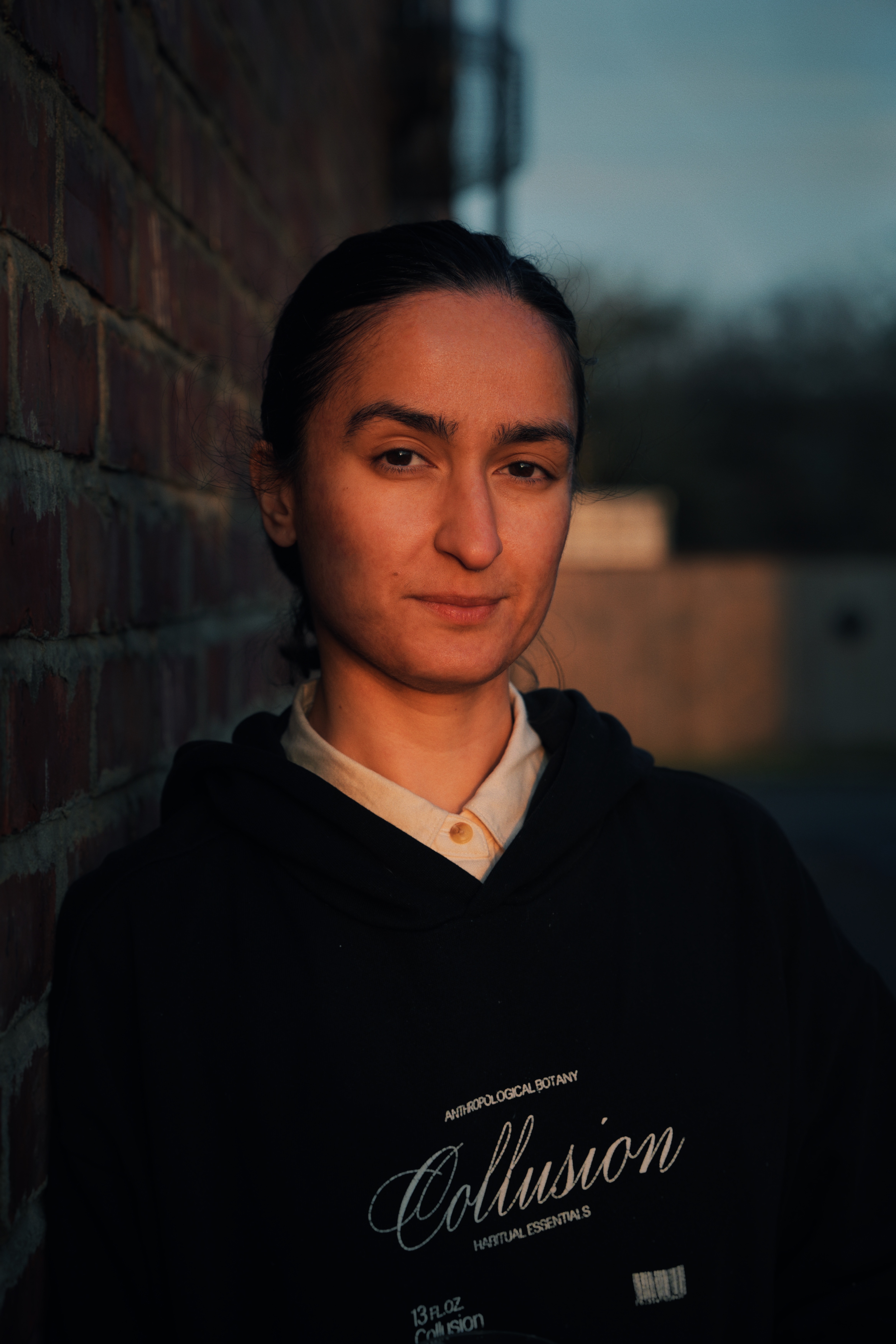 Hu-Sane stood by a brick wall and looking to camera lit by what could be a setting sun.