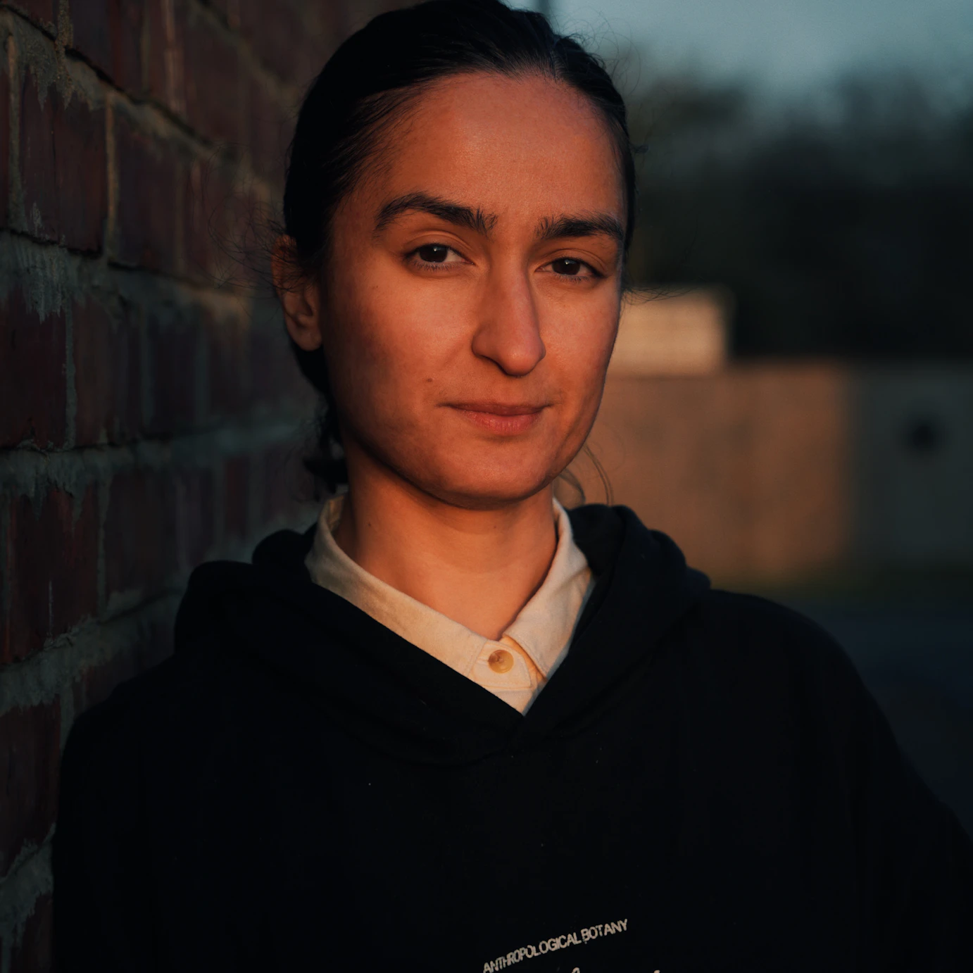 Hu-Sane stood by a brick wall and looking to camera lit by what could be a setting sun.
