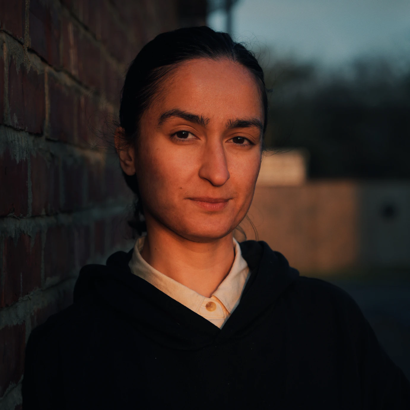 Hu-Sane stood by a brick wall and looking to camera lit by what could be a setting sun.