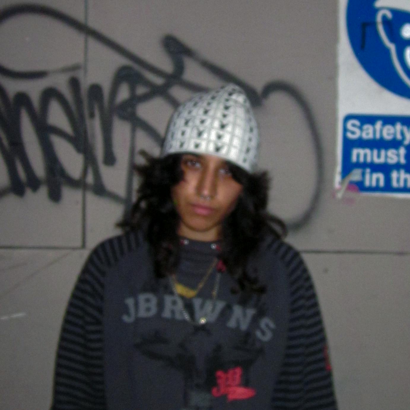 m3UNTITLED stood in front of a lightly graffitied wall looking to camera. The photo is slightly blurred. 