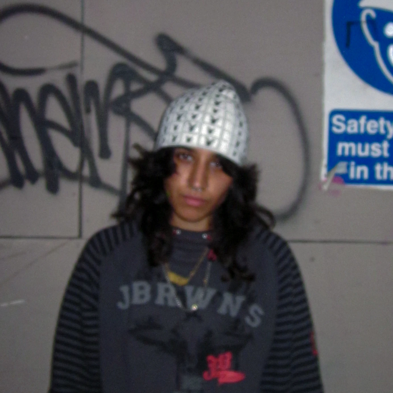 m3UNTITLED stood in front of a lightly graffitied wall looking to camera. The photo is slightly blurred.