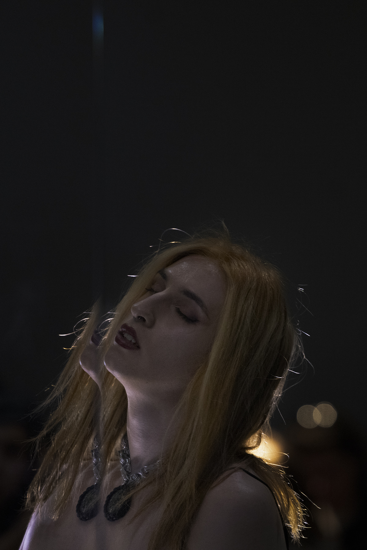 Phoenix Rousiamanis in a dark space, dimly lit from behind, facing slightly upwards with her eyes closed.