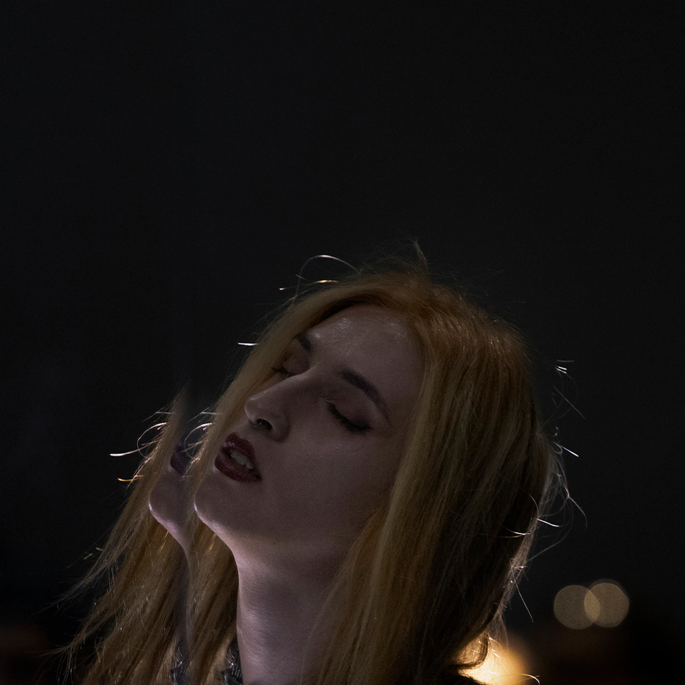 Phoenix Rousiamanis in a dark space, dimly lit from behind, facing slightly upwards with her eyes closed.