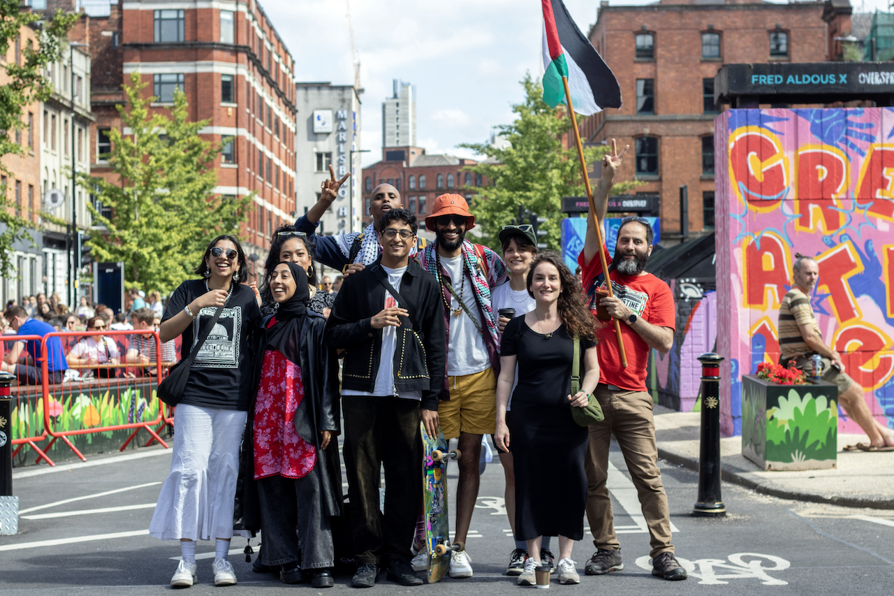 Member of MCR4PAL posing and smiling as a group in the street. One member on the right is holding a Palestinian flag on a pole.