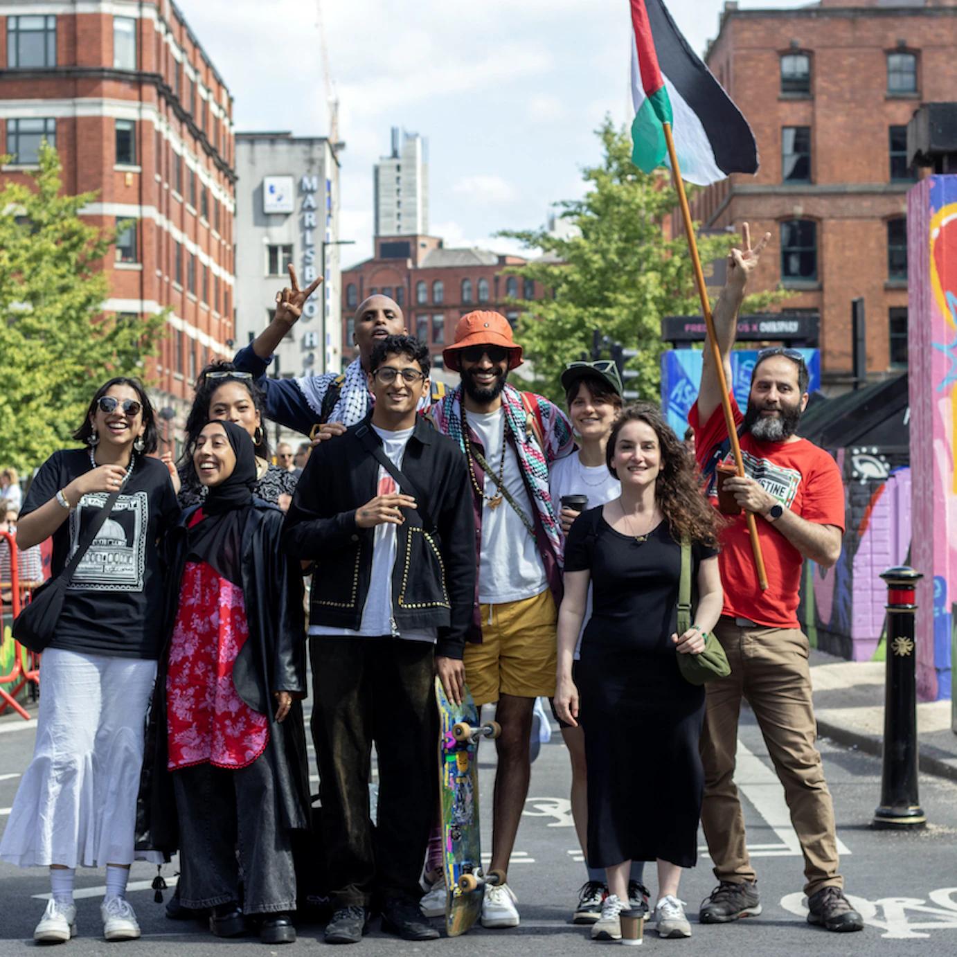 Member of MCR4PAL posing and smiling as a group in the street. One member on the right is holding a Palestinian flag on a pole.