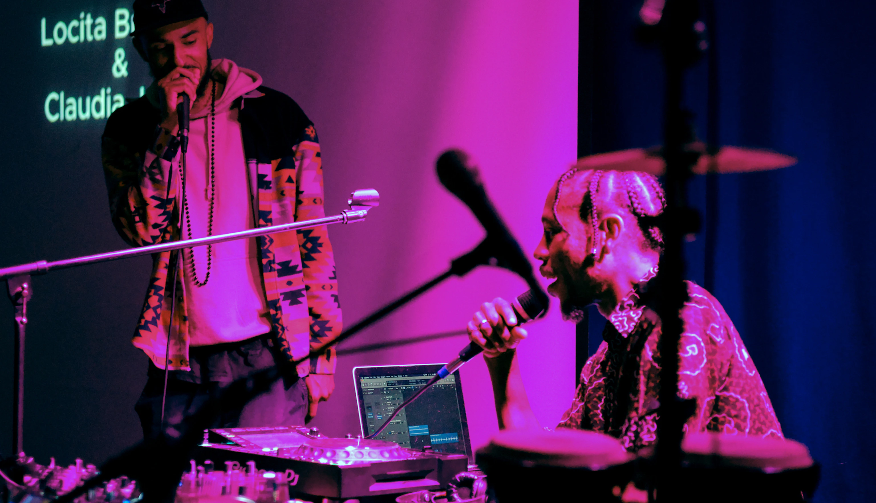 A musician sat by music production gear including a laptop, set of decks and microphones speaking to someone holding a microphone. The two are lit by pink lighting.