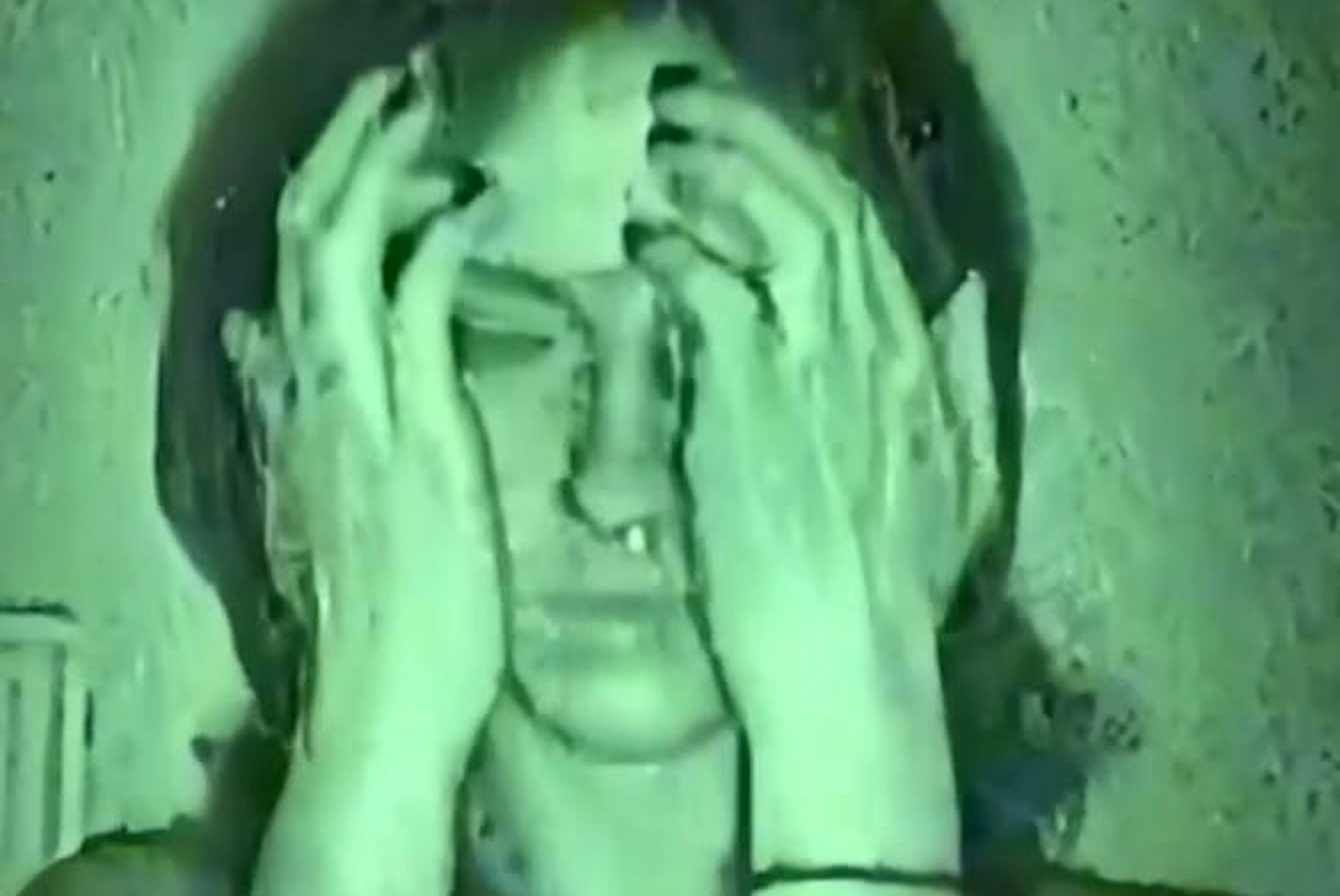 A still of Phoenix Rousiamanis being filmed via a night vision camcorder. Her hands are covering the sides of her head.