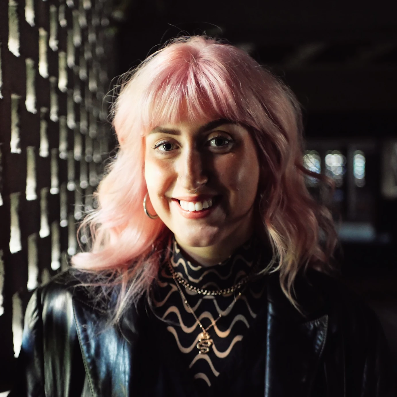 Sarah Barron stood by a stone wall with many square holes in it, shining sunlight through.